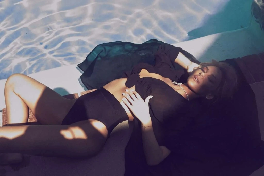 Katherine Taylor lying beside a swimming pool in sunlight, in a black swimsuit and an open black shirt