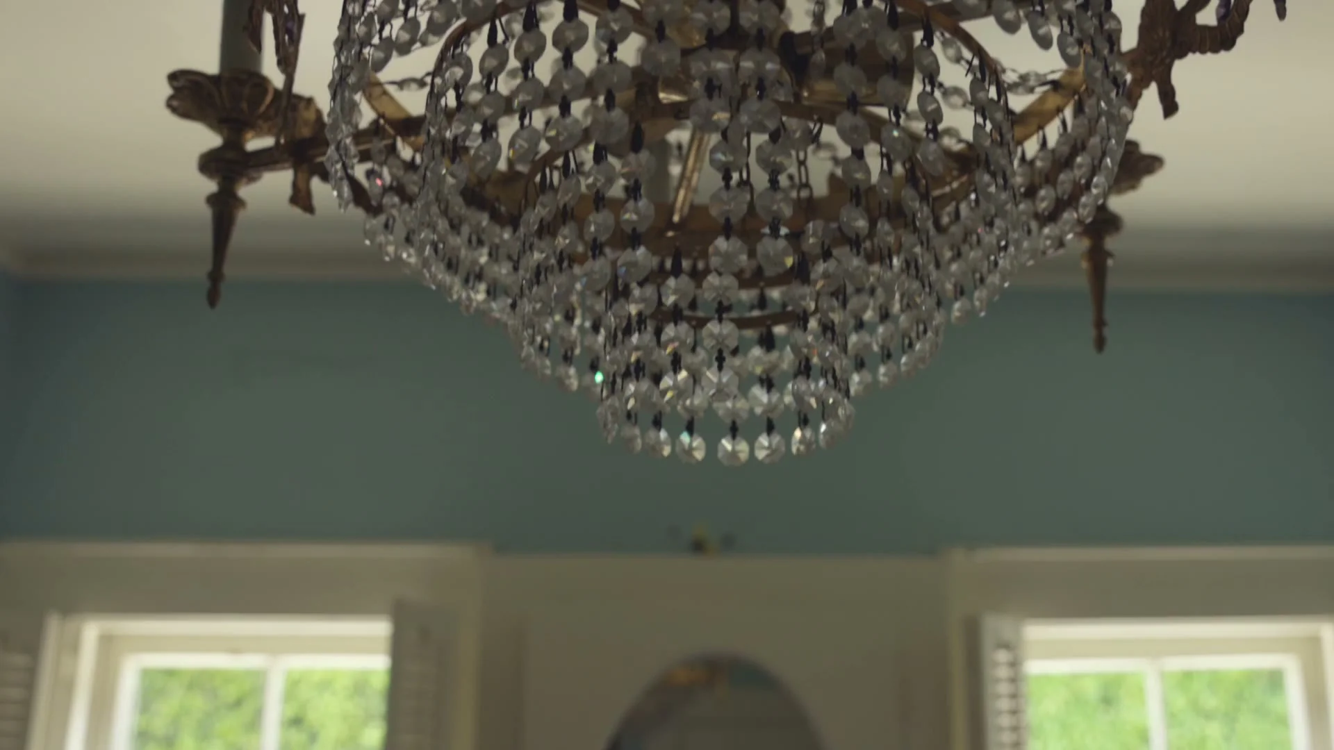 A crystal chandelier against a pale blue ceiling, with windows below