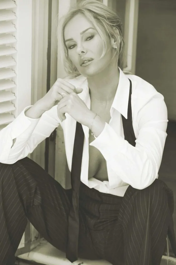 Katherine Taylor, independent escort and luxury companion, seated by white shutters in a white shirt, dark tie, and pinstripe trousers